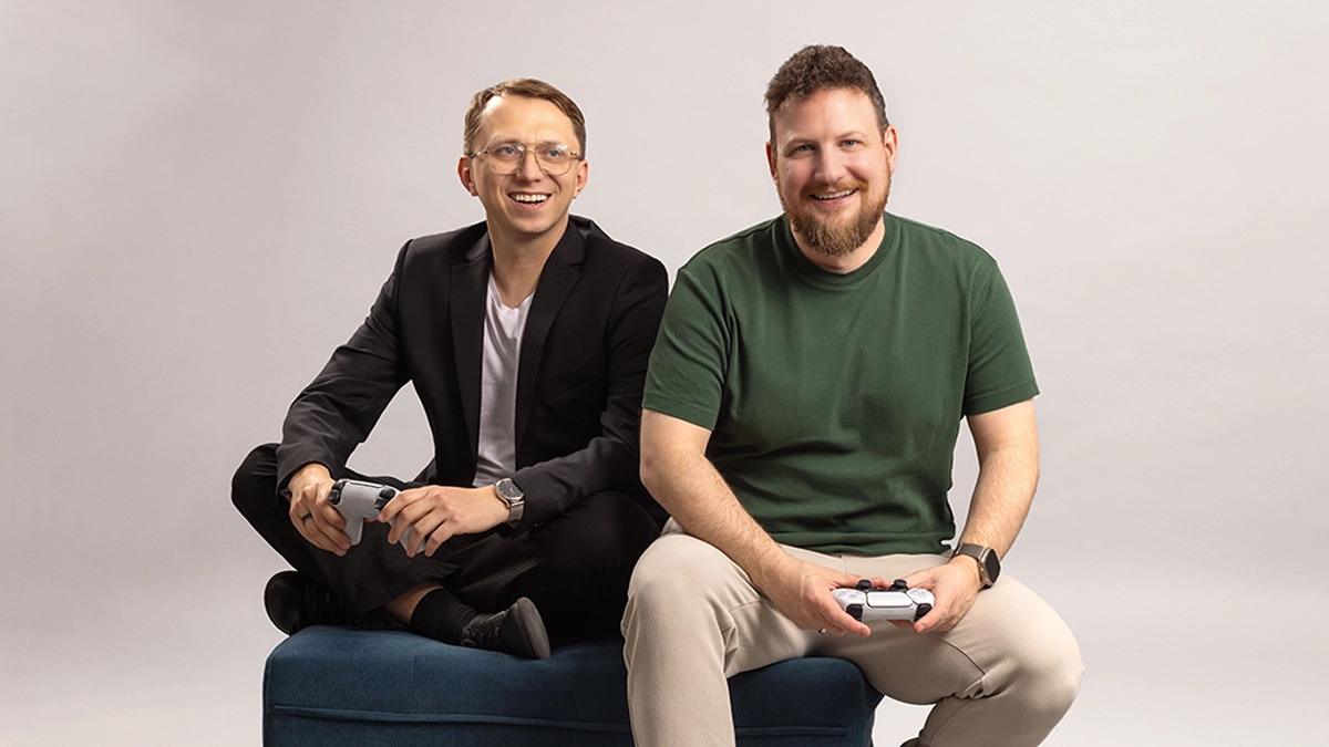 Two men sitting on a blue bench smiling, each holding a white gaming controller.