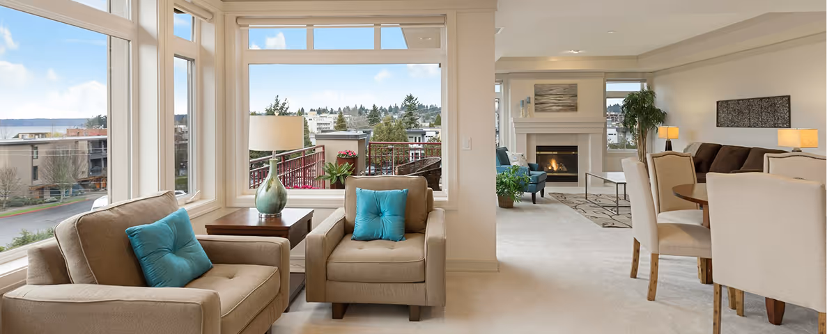 Bright living room with beige armchairs featuring turquoise cushions, large windows showing outdoor view, and a cozy fireplace in the background.