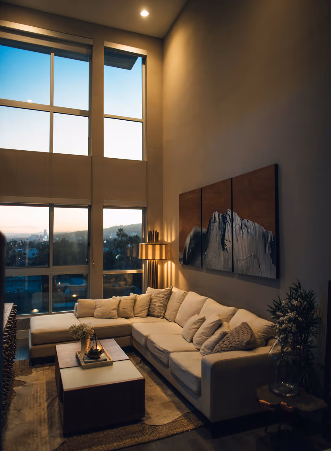 Cozy living room with a beige sectional sofa, coffee table, floor lamp, large windows, and mountain artwork on the wall.