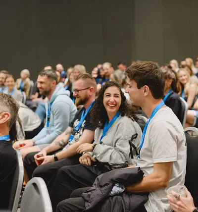 Testlio team sitting in rows of chairs at an event, with a woman smiling and engaging in conversation with a man beside her.