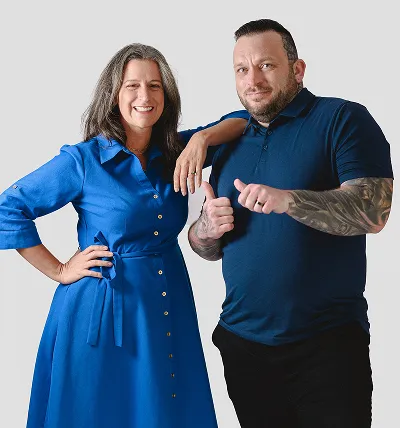 Smiling woman in a blue dress standing with her hand on her hip next to a man in a dark blue polo with tattoos giving a thumbs up.