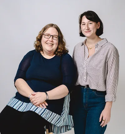 Testlio employees posing together against a plain light gray background; one seated wearing glasses and a navy blue dress with striped hem, the other standing wearing a striped button-up shirt and jeans.