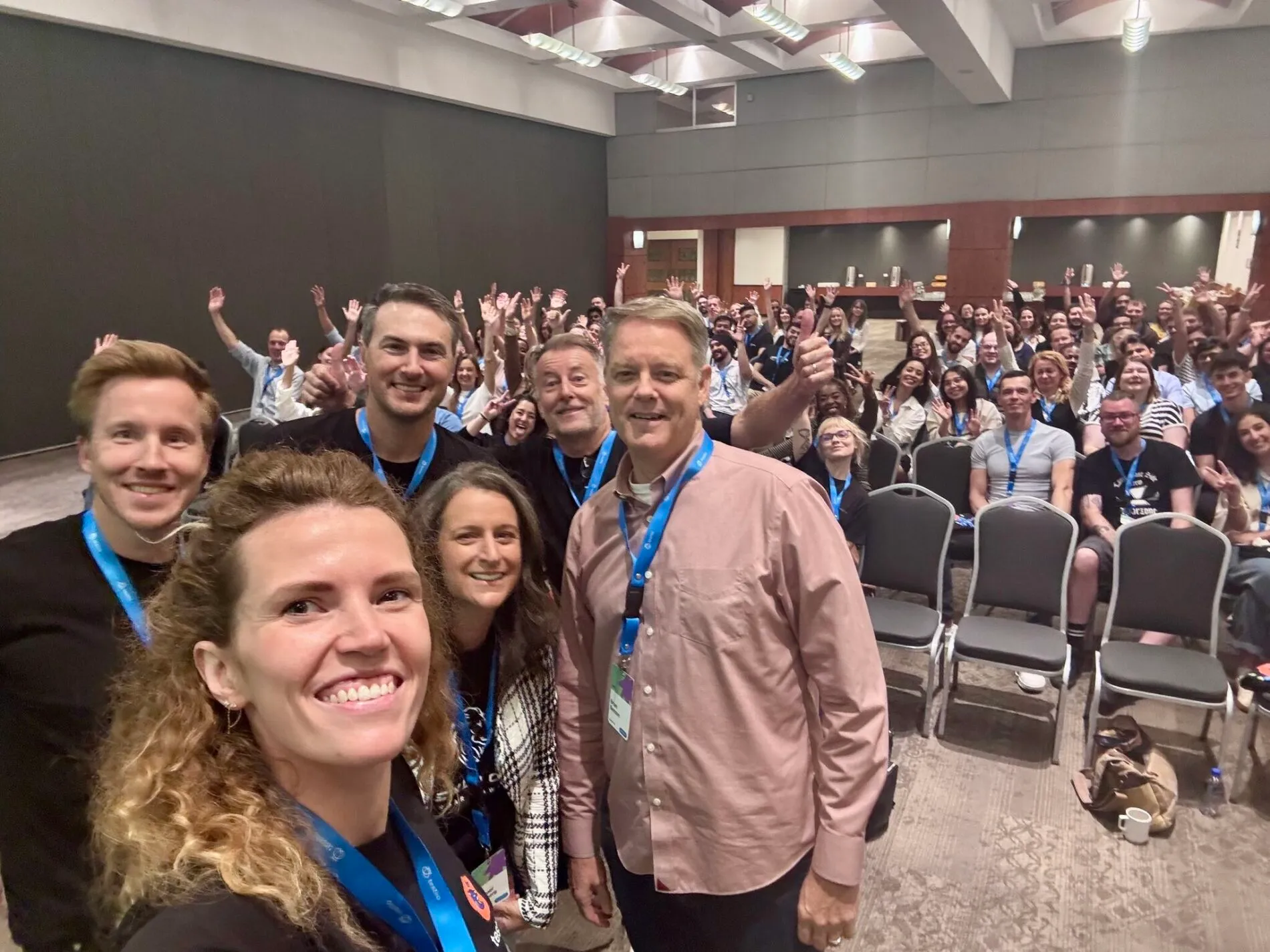 A large group of people from Testlio pose together in a conference room during LionFest. The photo is taken as a cheerful selfie from the front row, showing smiling team members and many people in the background waving and raising their hands.