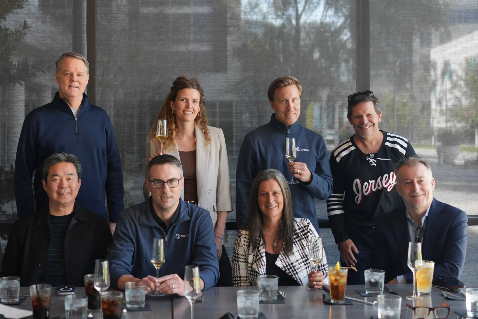 Group photo of Testlio’s board members and executive leadership team seated and standing around a restaurant table, some holding wine glasses. The group is arranged in two rows, with four people seated in front and four standing behind. From left to right, bottom row: Insik Rhee (Board Director), Mike Stearns (VP of Marketing), Summer Weisberg (CEO), and Dean Hickman-Smith (CRO). From left to right, top row: Brian Benson (CFO), Kristel Kruustük (Founder and Board Director), Marko Kruustük (Co-founder and Board Director), and Dave Keil (Board Director). Not pictured: Carmen Scarpa (Board Director) and Anthony Lee (Board Observer).