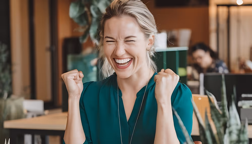 Smiling woman in green blouse cheering with clenched fists in an office setting.