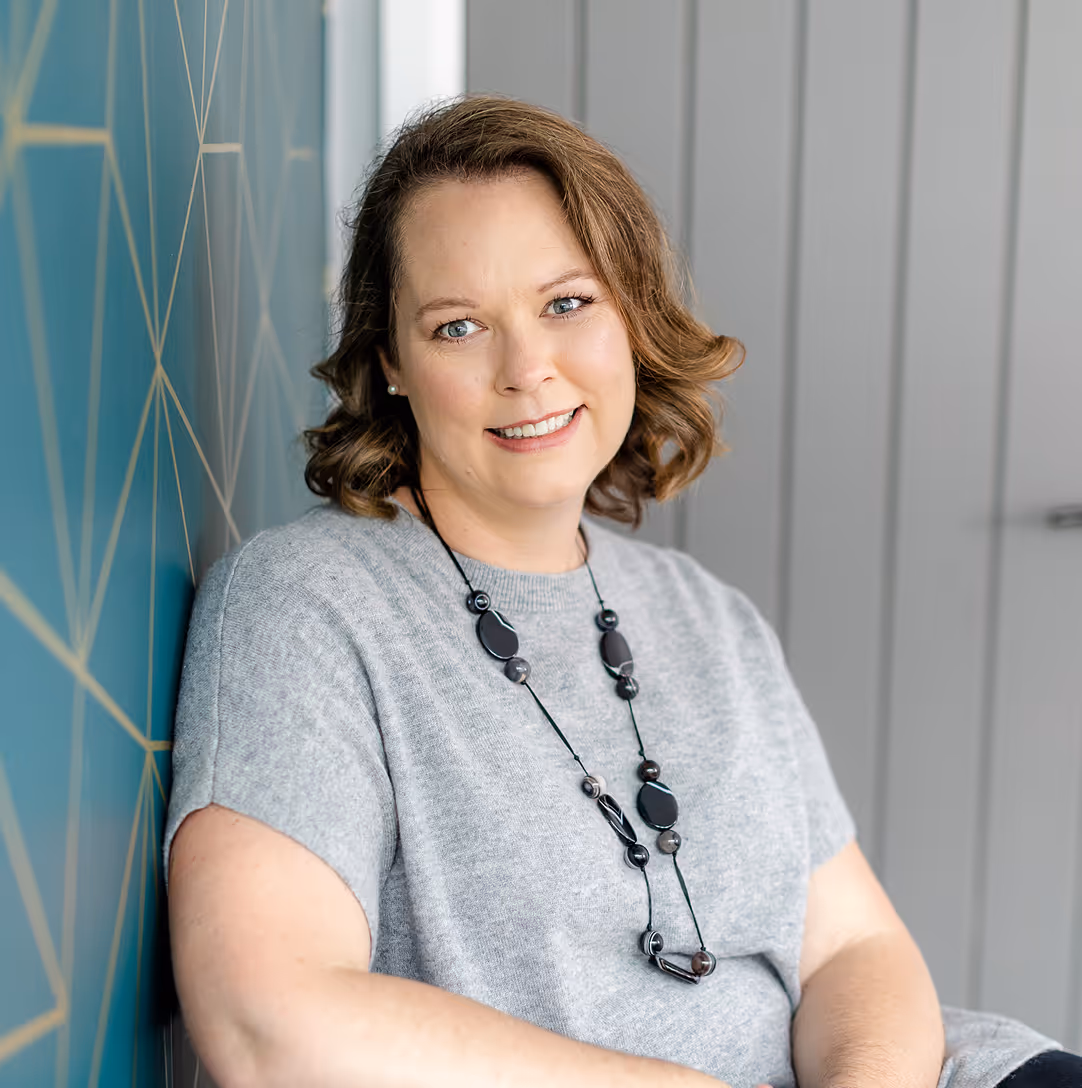Smiling woman with wavy brown hair wearing a gray short-sleeve sweater and a black beaded necklace, sitting against a blue geometric patterned wall.
