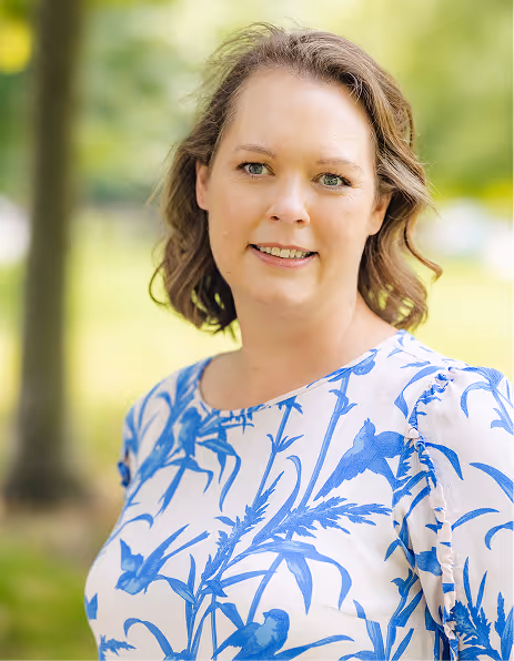 Smiling woman with shoulder-length brown hair wearing a white blouse with blue bird and branch patterns outdoors with blurred greenery background.
