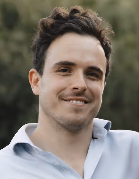 Smiling man with curly hair wearing a light blue shirt outdoors.