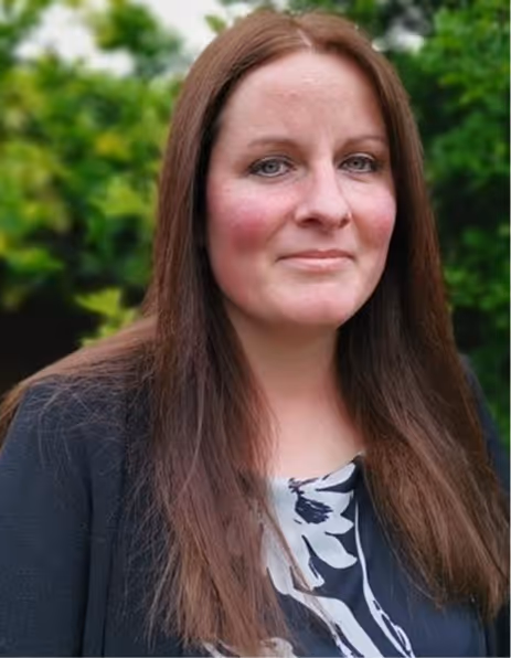 Woman with long brown hair wearing a black floral top and black jacket, posing outdoors with greenery in the background.