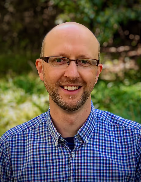Smiling bald man with glasses wearing a blue and white checkered shirt outdoors with green foliage background.