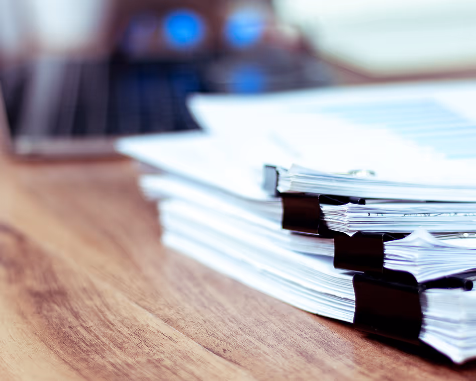 Stack of paper documents held by black binder clips on a wooden desk with a blurred laptop in the background.