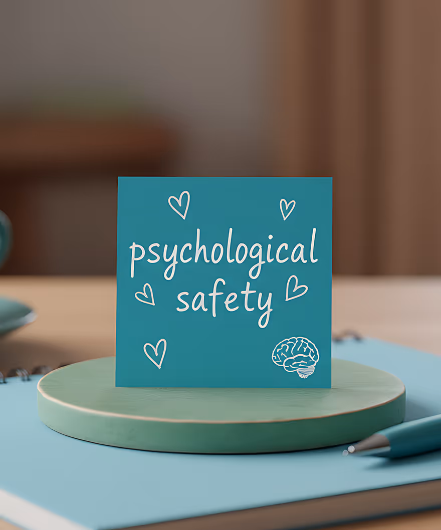 Blue card with the words 'psychological safety' surrounded by heart doodles and a brain illustration, standing on a round green platform on a desk.