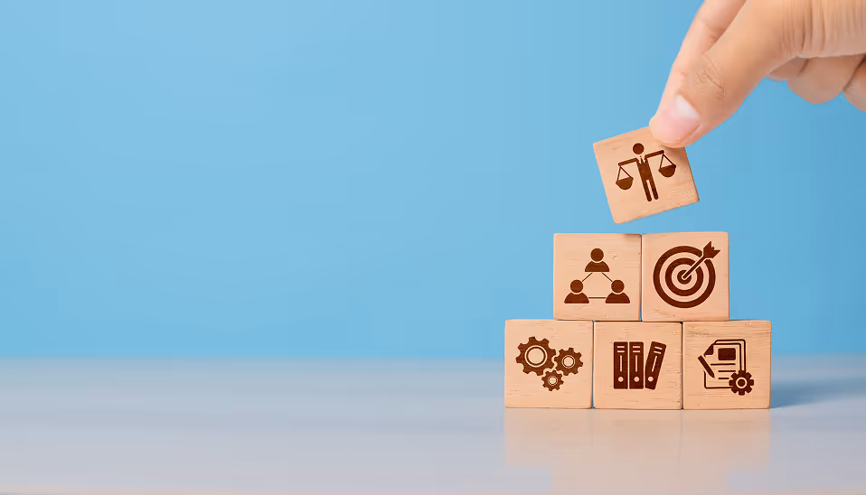 Hand placing a wooden block with a justice scale icon on top of a pyramid of blocks showing icons for teamwork, target, gears, files, and document settings.