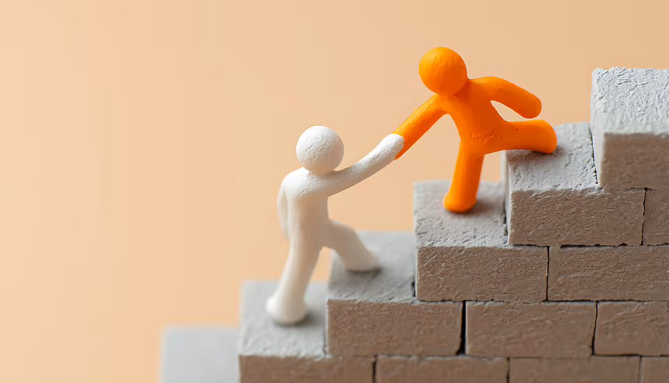 Orange figure helping white figure climb gray stone stairs, symbolizing support and teamwork.