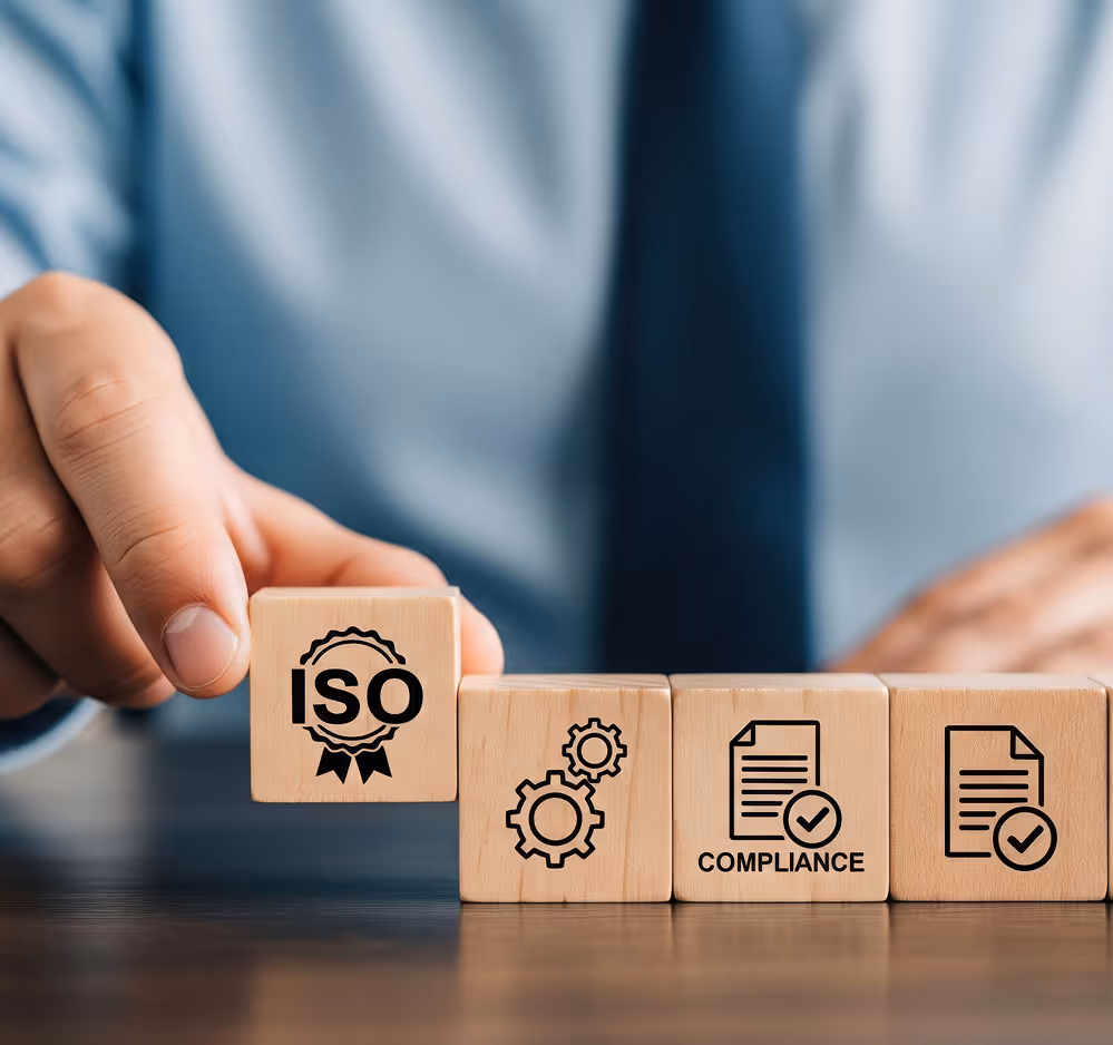 Hand placing a wooden block with ISO text and badge icon next to other blocks showing gears and compliance documents with checkmarks.