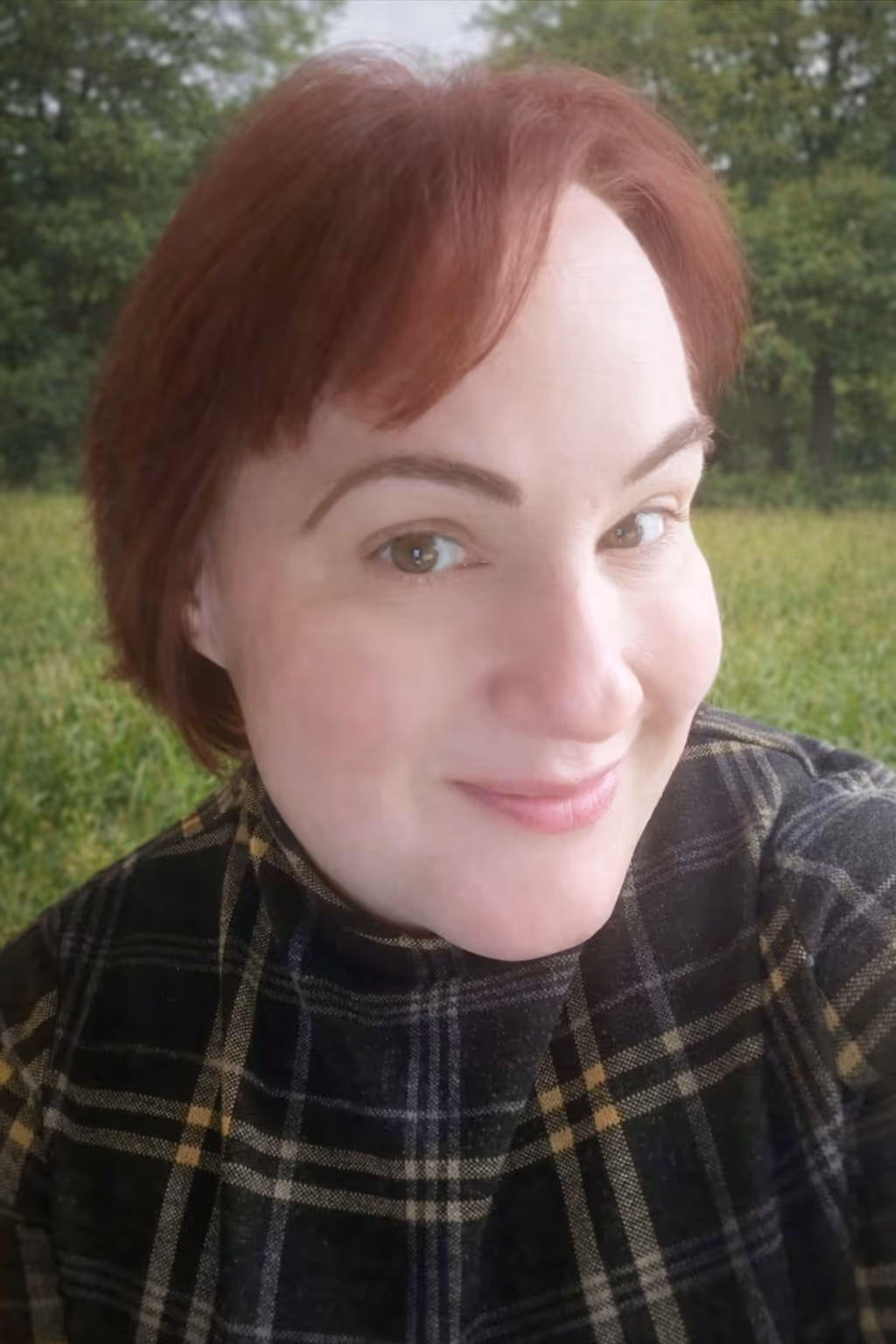 Smiling woman with short red hair wearing a black and yellow plaid shirt outdoors with green trees and grass in the background.