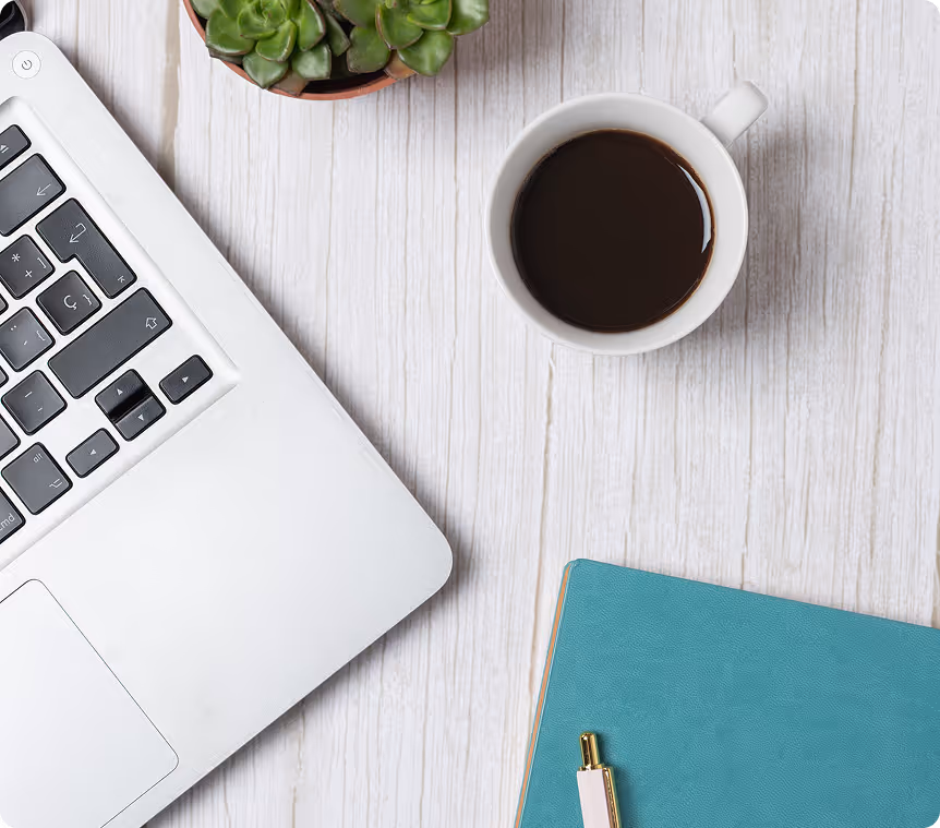 Top view of a workspace with a silver laptop, a cup of black coffee, a small green succulent plant, and a teal notebook with a pen on a white wooden surface.