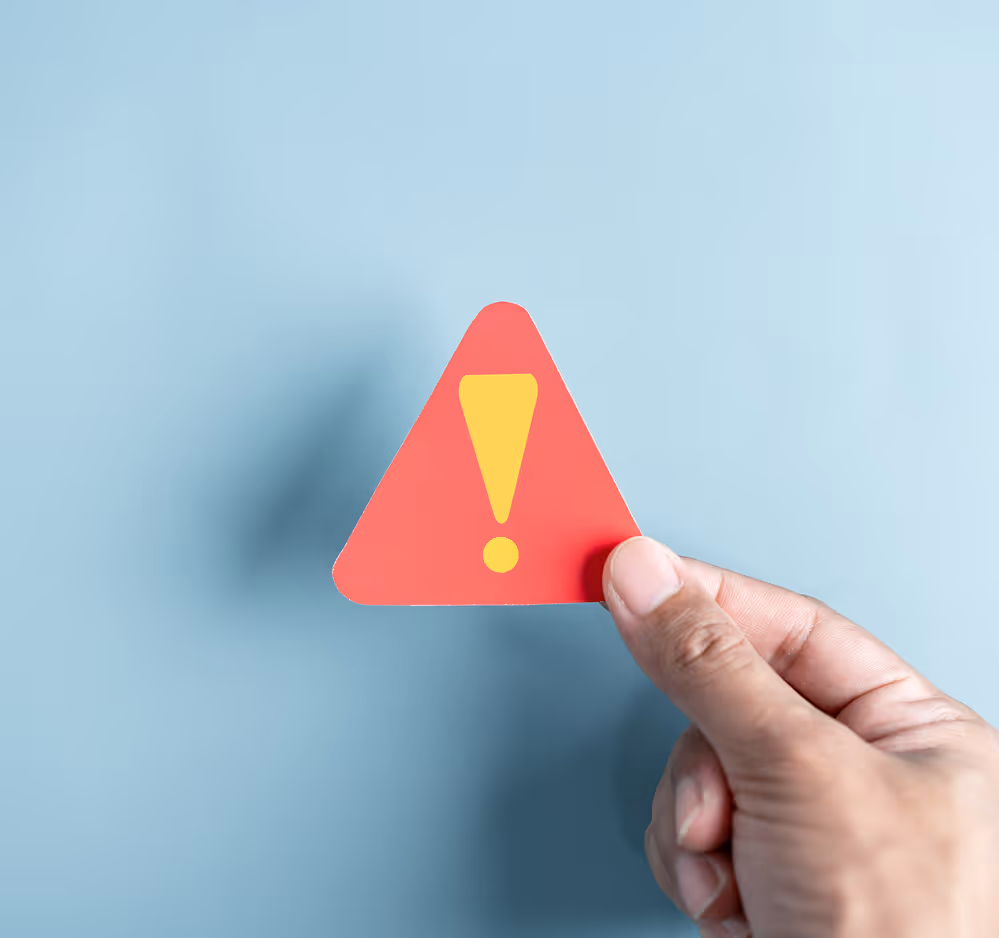 Hand holding a red triangular warning sign with a yellow exclamation mark against a blue background.