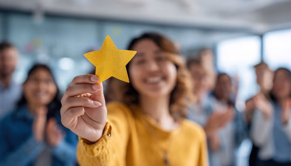 Smiling woman in yellow holding a gold star with blurred people clapping in the background.