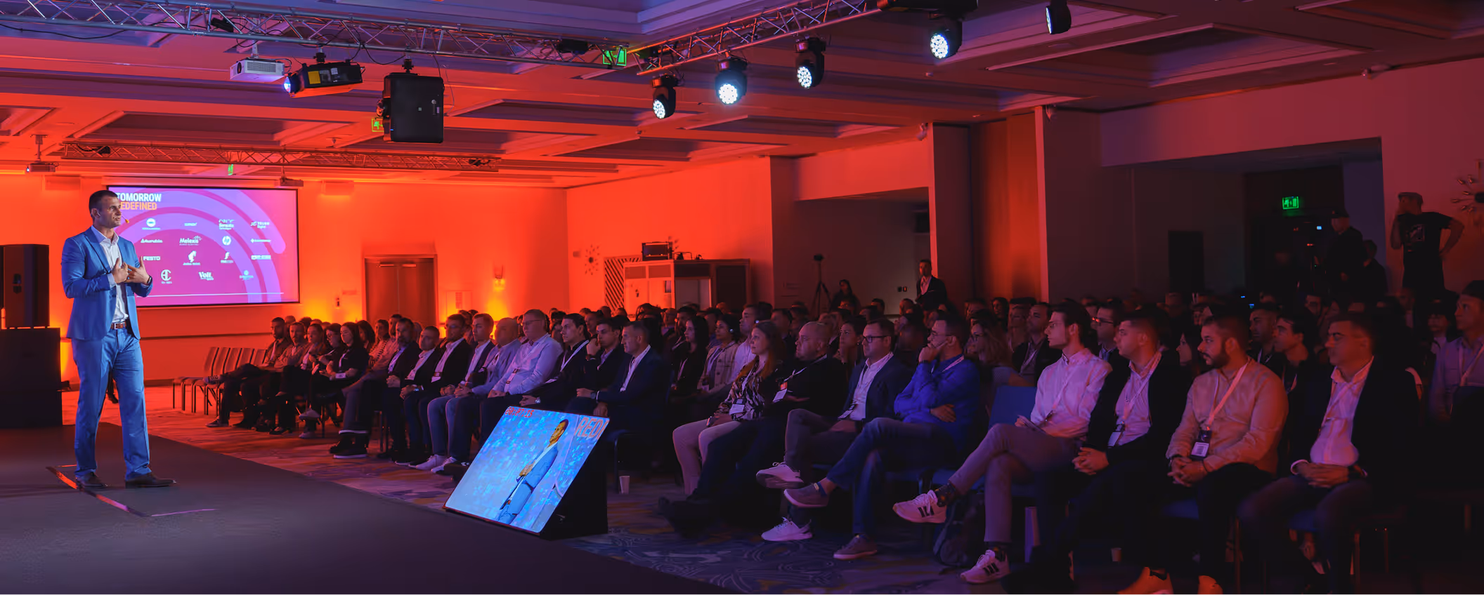 Man in blue suit giving a presentation on stage to a seated audience in a dimly lit conference room with an orange-red background.