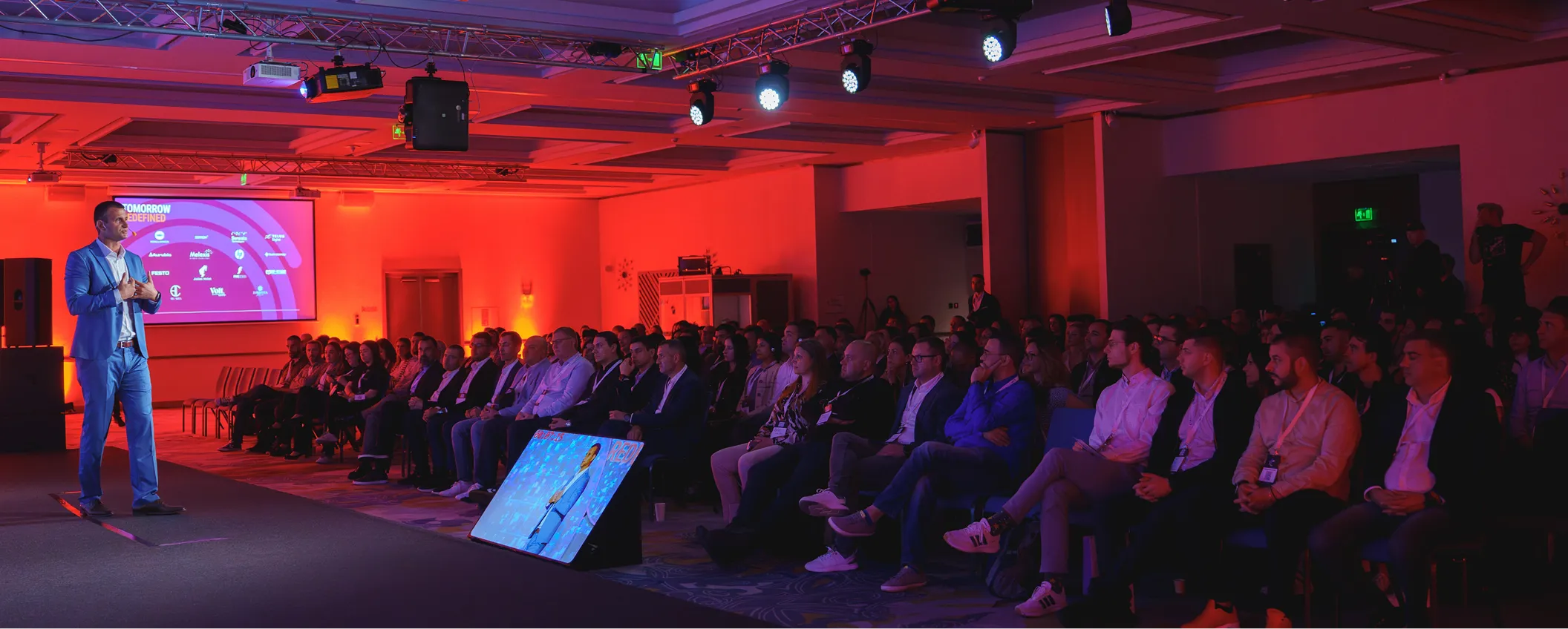 Man in blue suit giving a presentation on stage to a seated audience in a dimly lit conference room with an orange-red background.