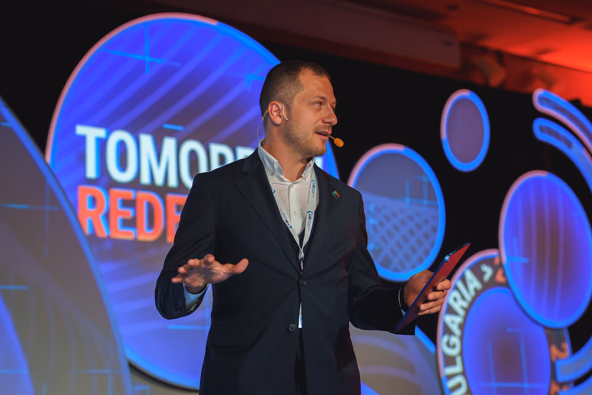 Man in dark suit speaking on stage with a microphone headset and holding a tablet, with colorful circular digital graphics in the background.