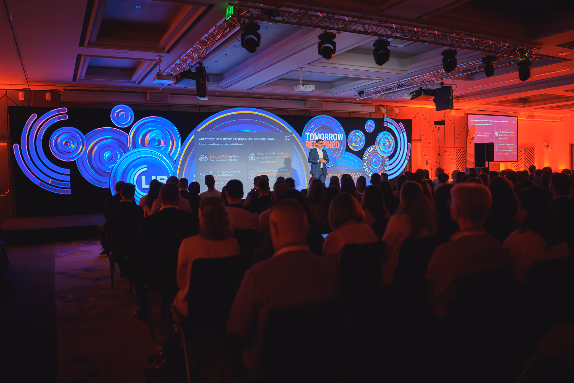 Audience watching a speaker on stage at a conference with illuminated blue circular graphics and text reading 'Tomorrow Reinvented'.