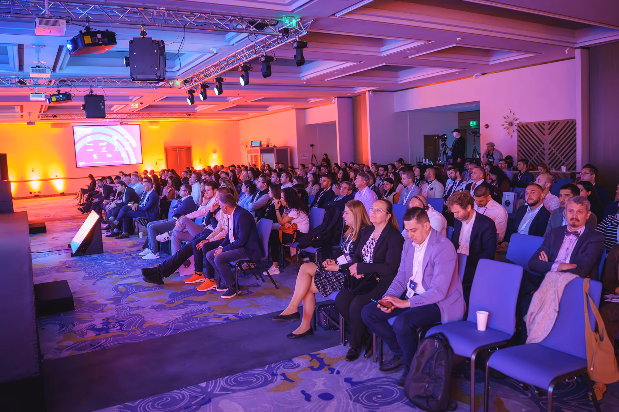 Audience seated attentively in a conference room with purple and orange lighting during a presentation.
