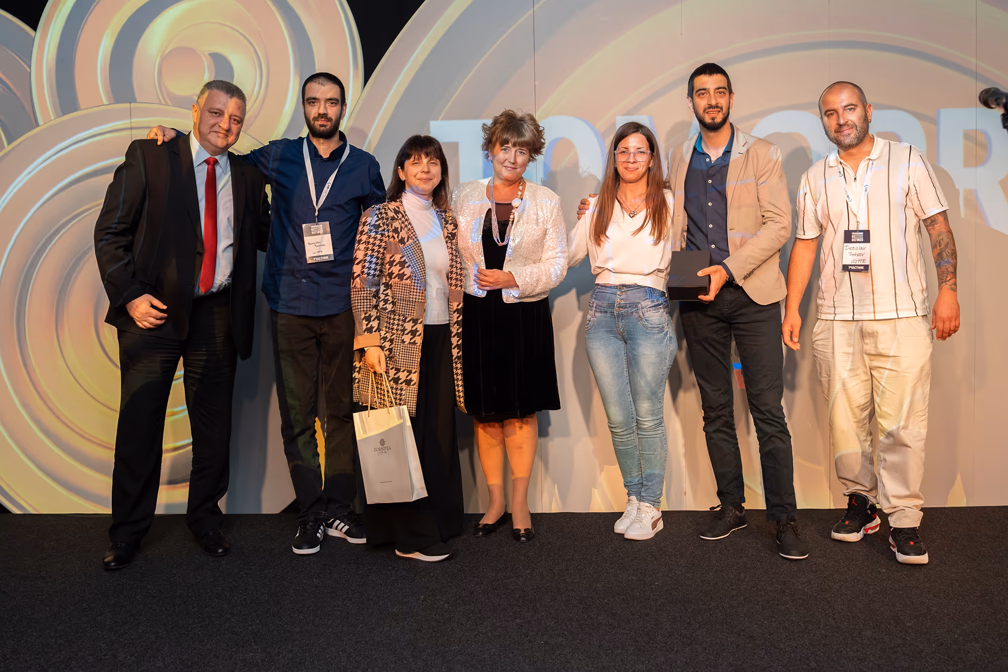 Group of seven people standing on a stage with circular light patterns in the background, some holding awards and smiling.
