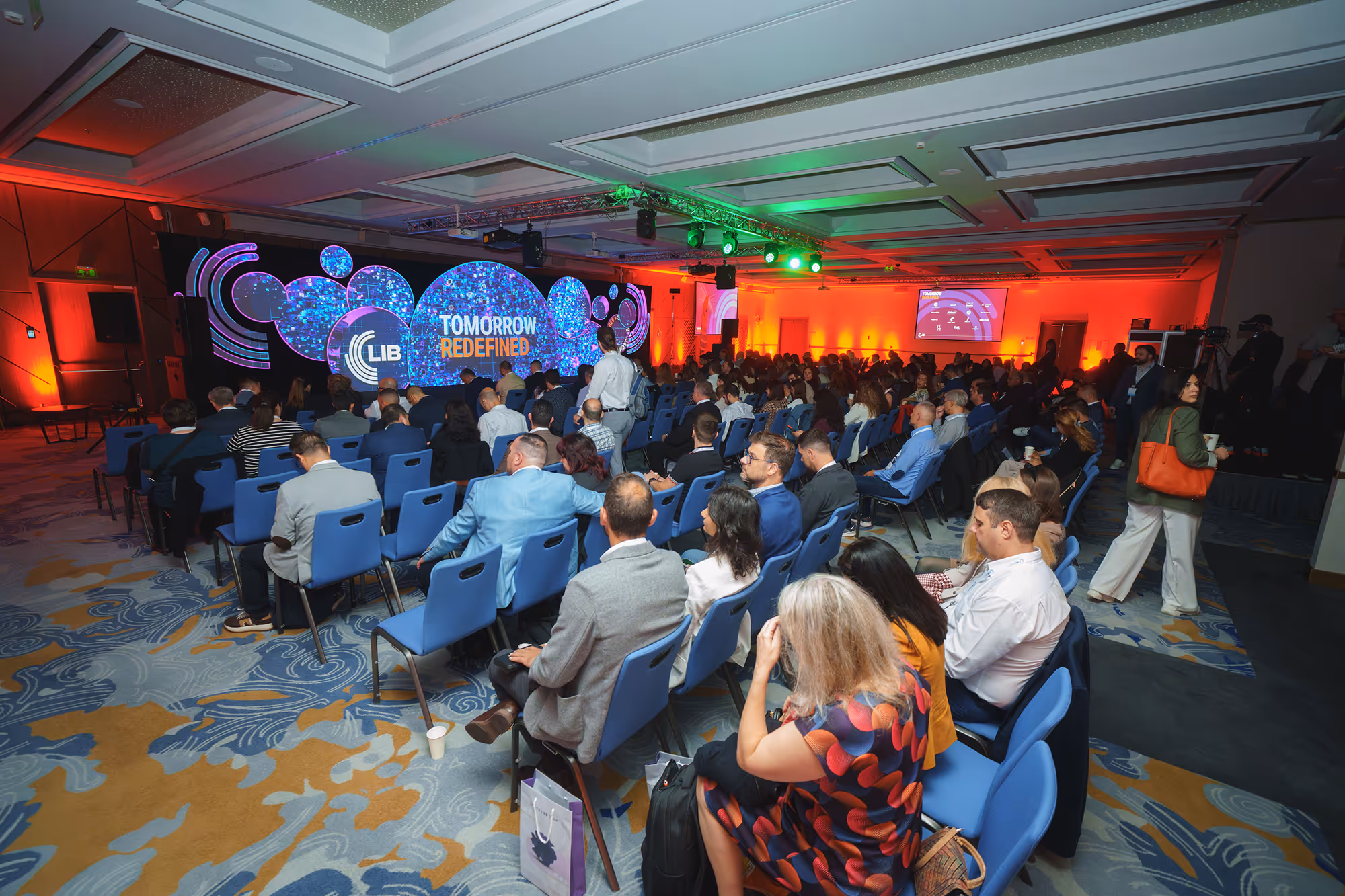 Audience seated in a conference room facing a large stage with colorful digital screens displaying 'LIB TOMORROW REDEFINED'.
