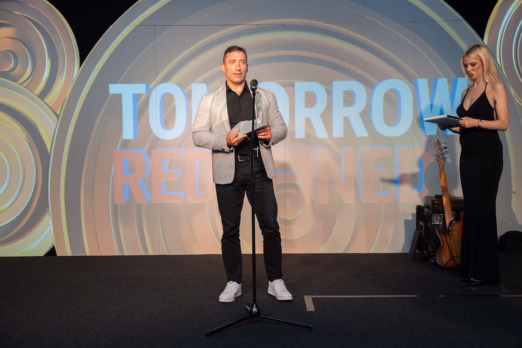 Man in a gray blazer and black shirt speaking on stage with a microphone, a woman in a black dress stands nearby reading from a notebook.