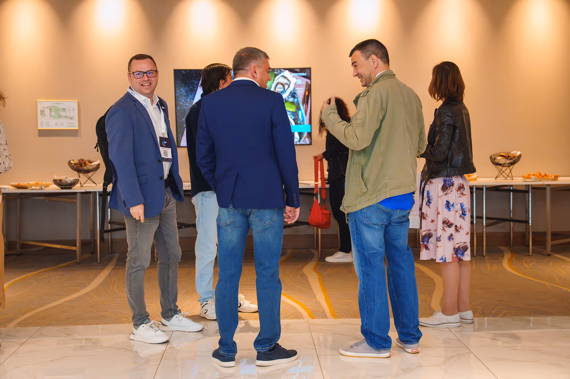 People in casual and business attire chatting in a brightly lit indoor space with tables of snacks and a screen in the background.