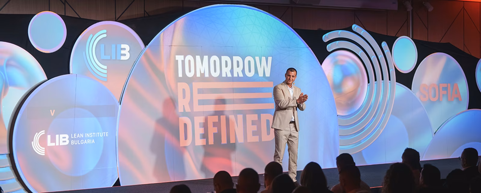 Лектор изнася презентация на модерната брандирана сцена с логото на Lean Institute Bulgaria и Tomorrow Redefined.