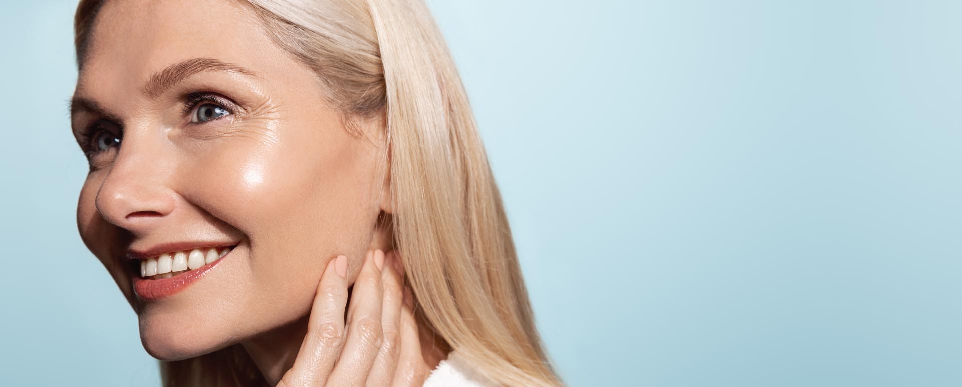 Nærbilde av en smilende middelaldrende kvinne med langt blondt hår og glødende hud mot en blå bakgrunn.