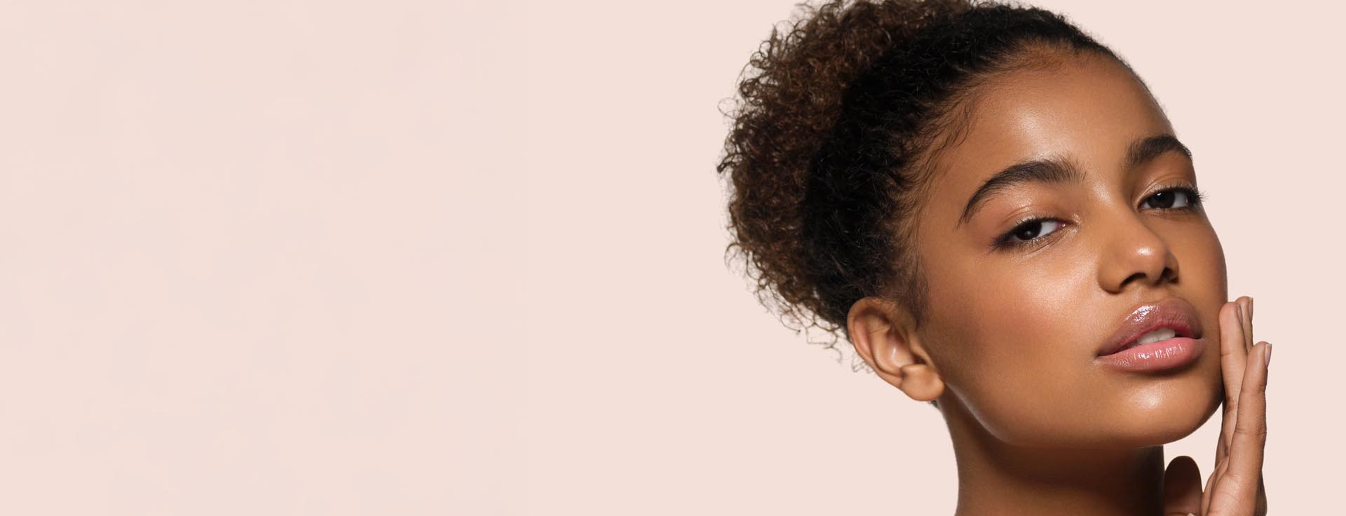 Nærbilde av kvinne med krøllete hår som berører kinnet sitt med hånden mot lys bakgrunn.