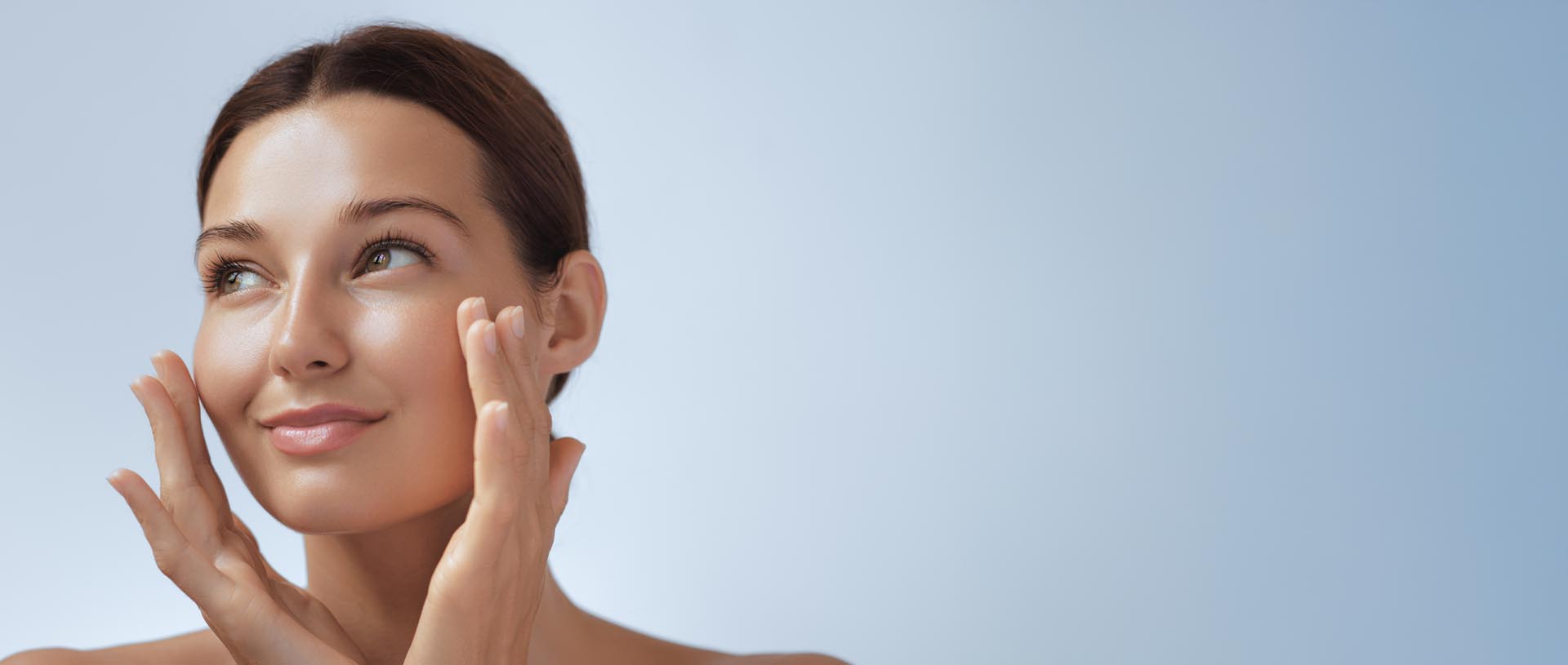 Kvinne med glatt, sunn hud berører ansiktet forsiktig med begge hender og smiler svakt.