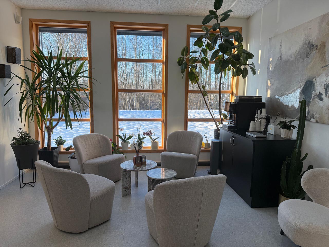 Moderne sittegruppe med fire beige stoler rundt to små marmorbord i et lyst rom med store vinduer, grønne planter og et svart skap med kaffemaskin.