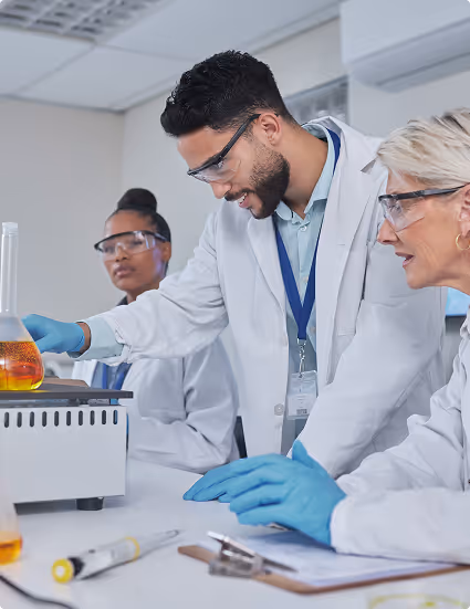 Three scientists wearing lab coats, safety glasses, and gloves working with laboratory glassware and equipment.