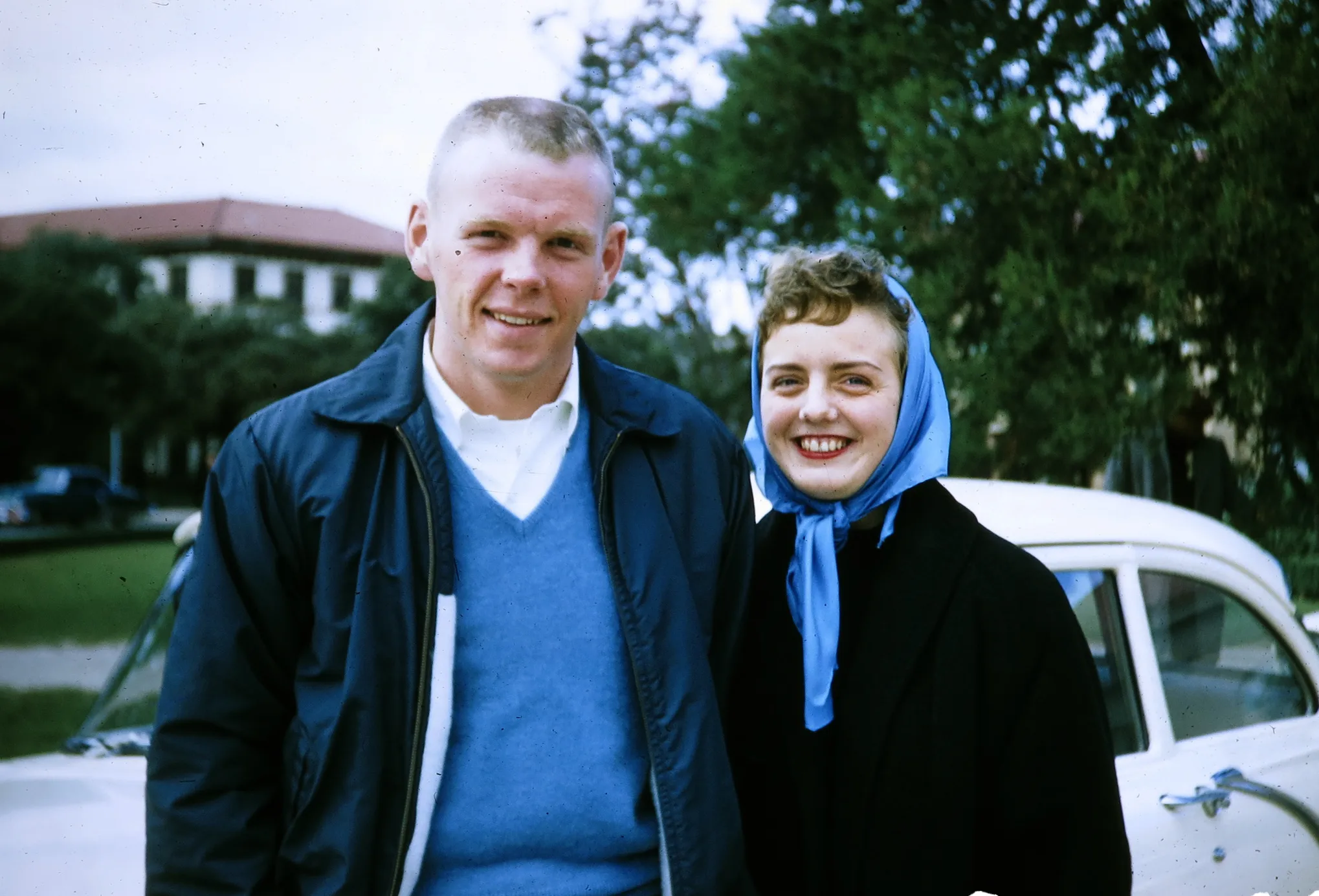 Smiling young man in a blue sweater and jacket standing next to a woman wearing a blue headscarf and black coat in front of a white car.