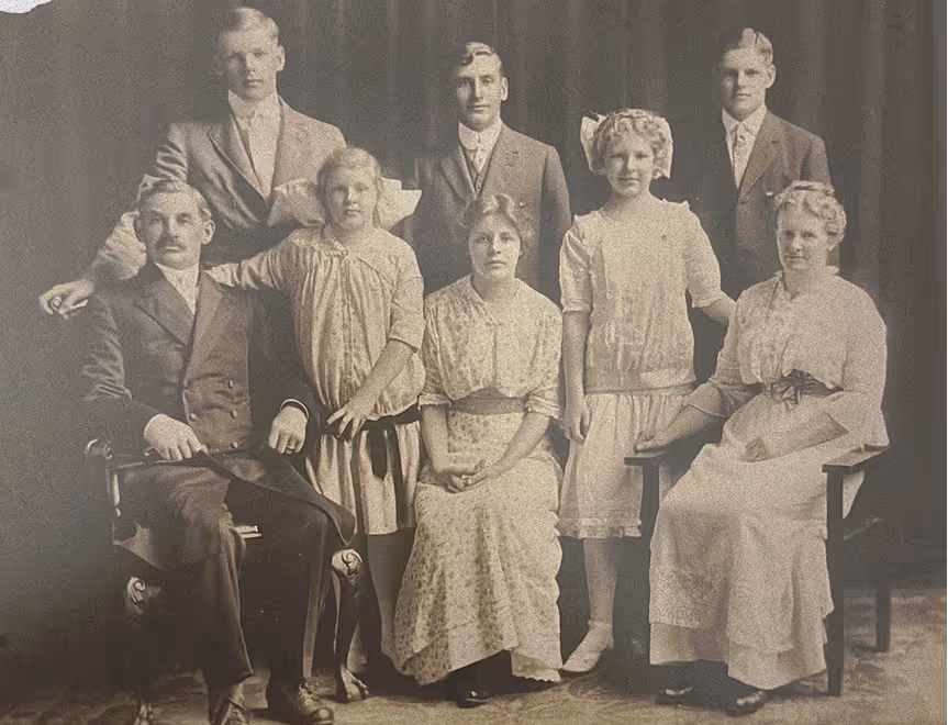 Sepia-toned vintage photo of eight formally dressed people, five women and three men, posing indoors.