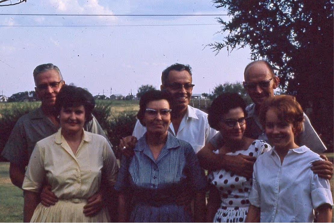 Group of seven adults standing closely together outside on a grassy area with trees and a clear sky in the background.