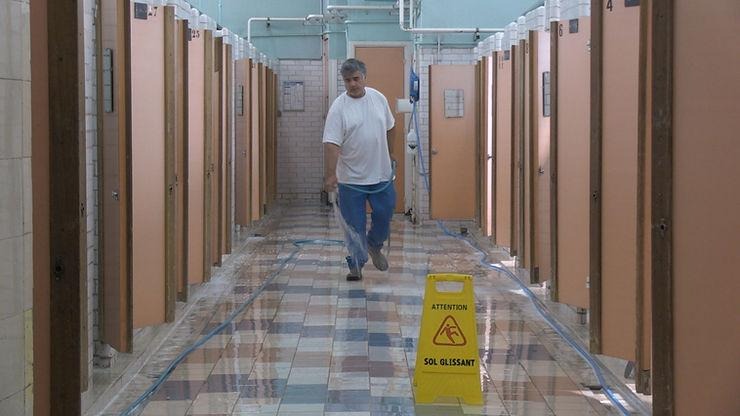 Cette photo en couleur représente un employé des bains douches qui lave le sol. Les cabines sont de chaque coté. A droite en bas de l'image, un panneau jaune indique que le sol est glissant. 