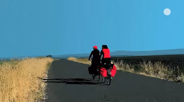 Illustration numérique au style graphique épuré représentant deux personnes voyageant à vélo sur une route de campagne. Vues de dos, elles roulent côte à côte sur des vélos équipés de sacoches de voyage rouges, au milieu d’un paysage ouvert bordé de champs. L’une porte une casquette rouge, l’autre un haut rouge vif, créant un contraste marqué avec le ciel bleu uniforme. La route s’étire vers l’horizon, renforçant l’impression de liberté, d’aventure et de long voyage. Cette image évoque le cyclotourisme, l’entraide et le dépassement de soi. Copyright Cortex Média.