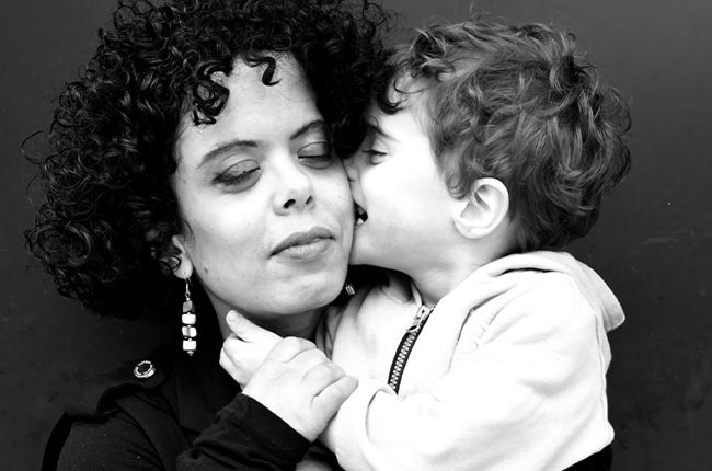 Femme aux cheveux bouclés tenant un jeune enfant dans ses bras ; l’enfant lui embrasse la joue. Portrait en noir et blanc sur fond uni, exprimant tendresse et proximité.