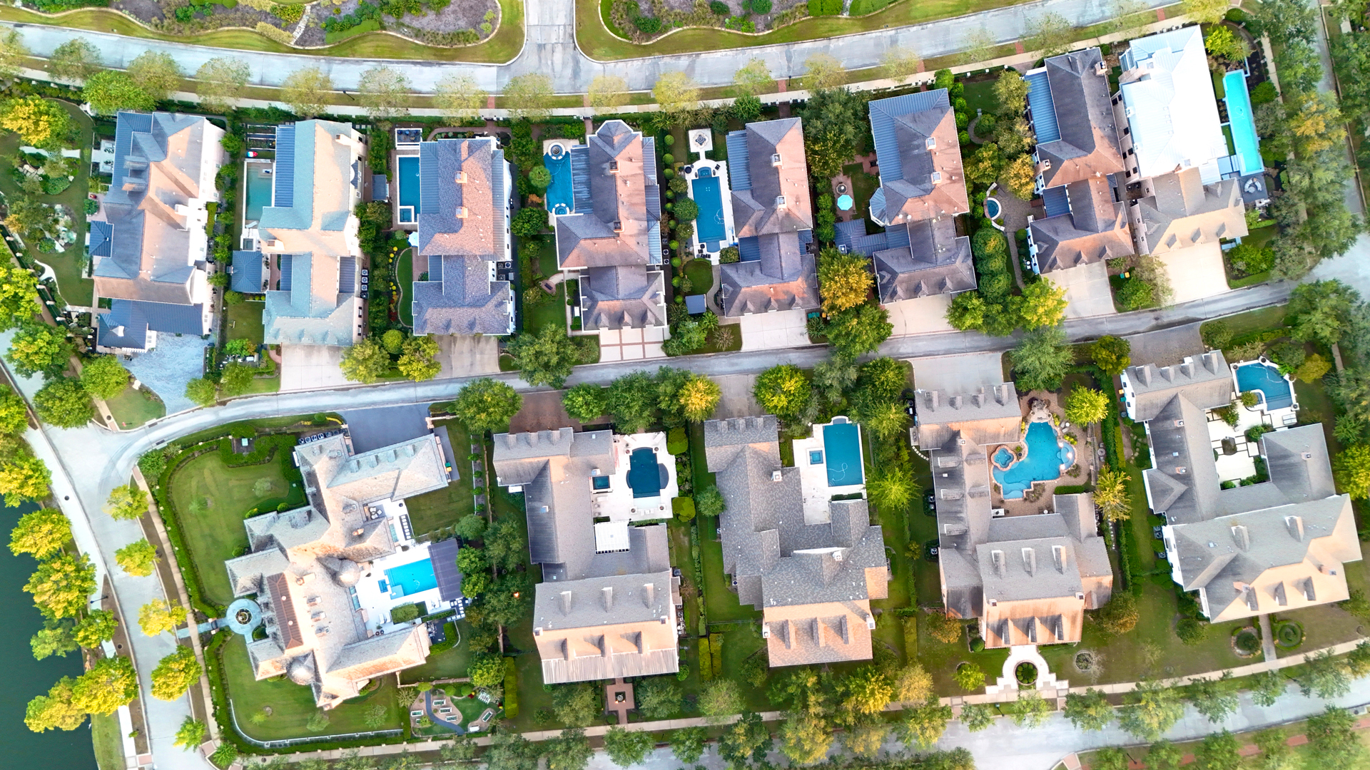 Aerial view of a suburban neighborhood with large houses, green lawns, and several swimming pools.
