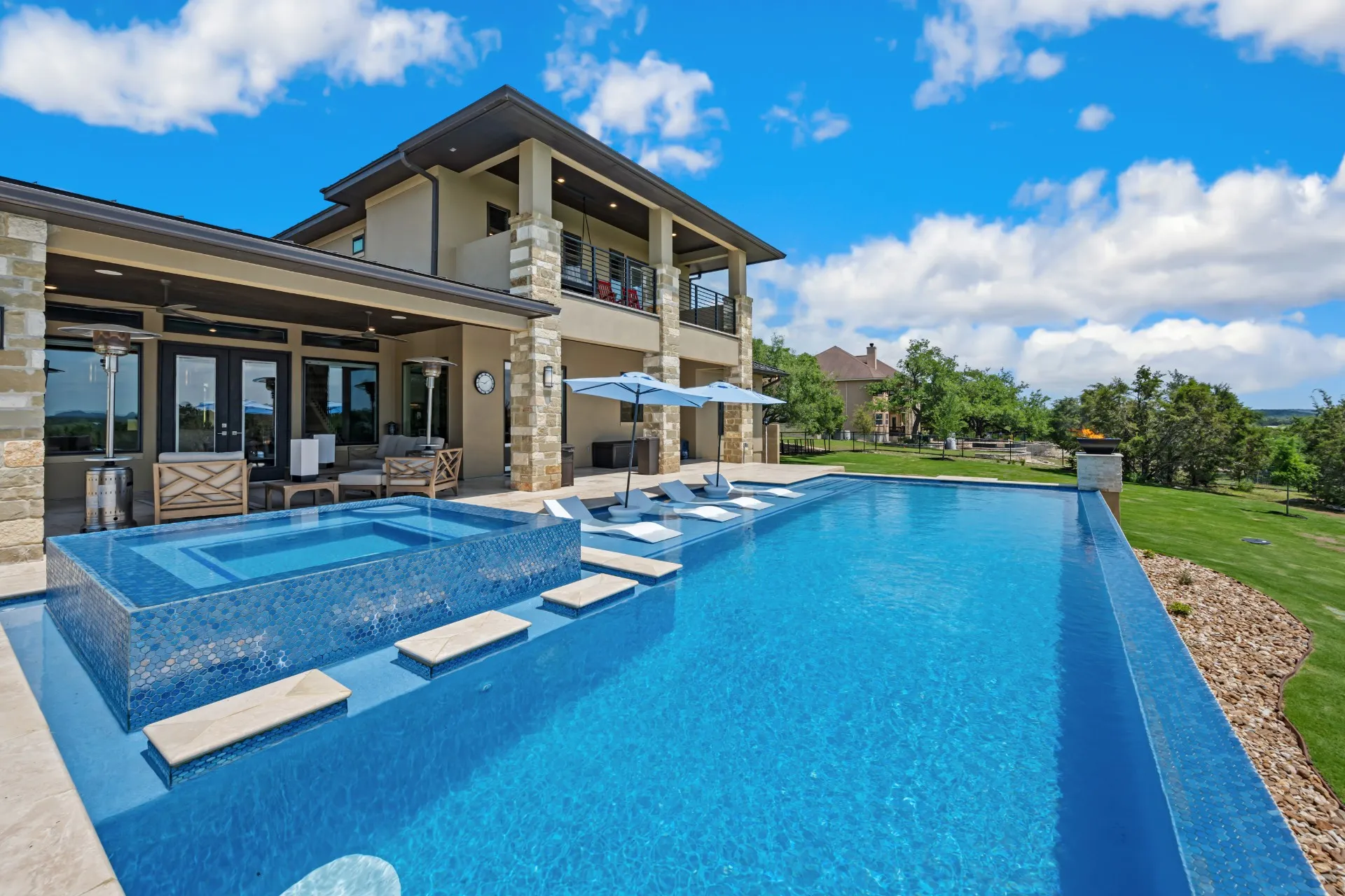 Modern two-story house with an outdoor infinity pool featuring built-in loungers and umbrellas under a bright blue sky.