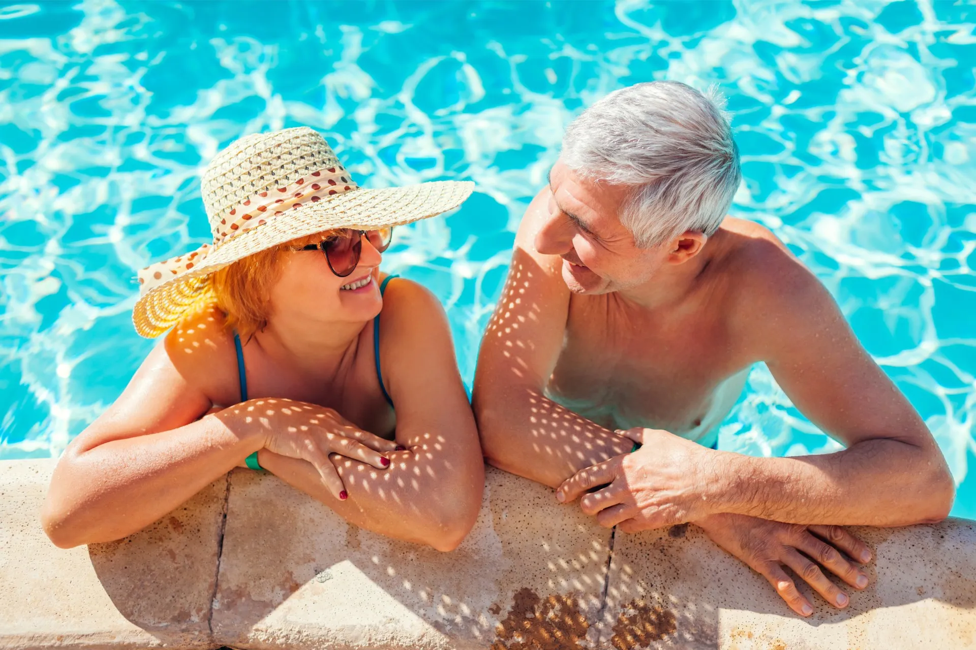 Smiling elderly couple leaning on the edge of a pool, enjoying a sunny day.