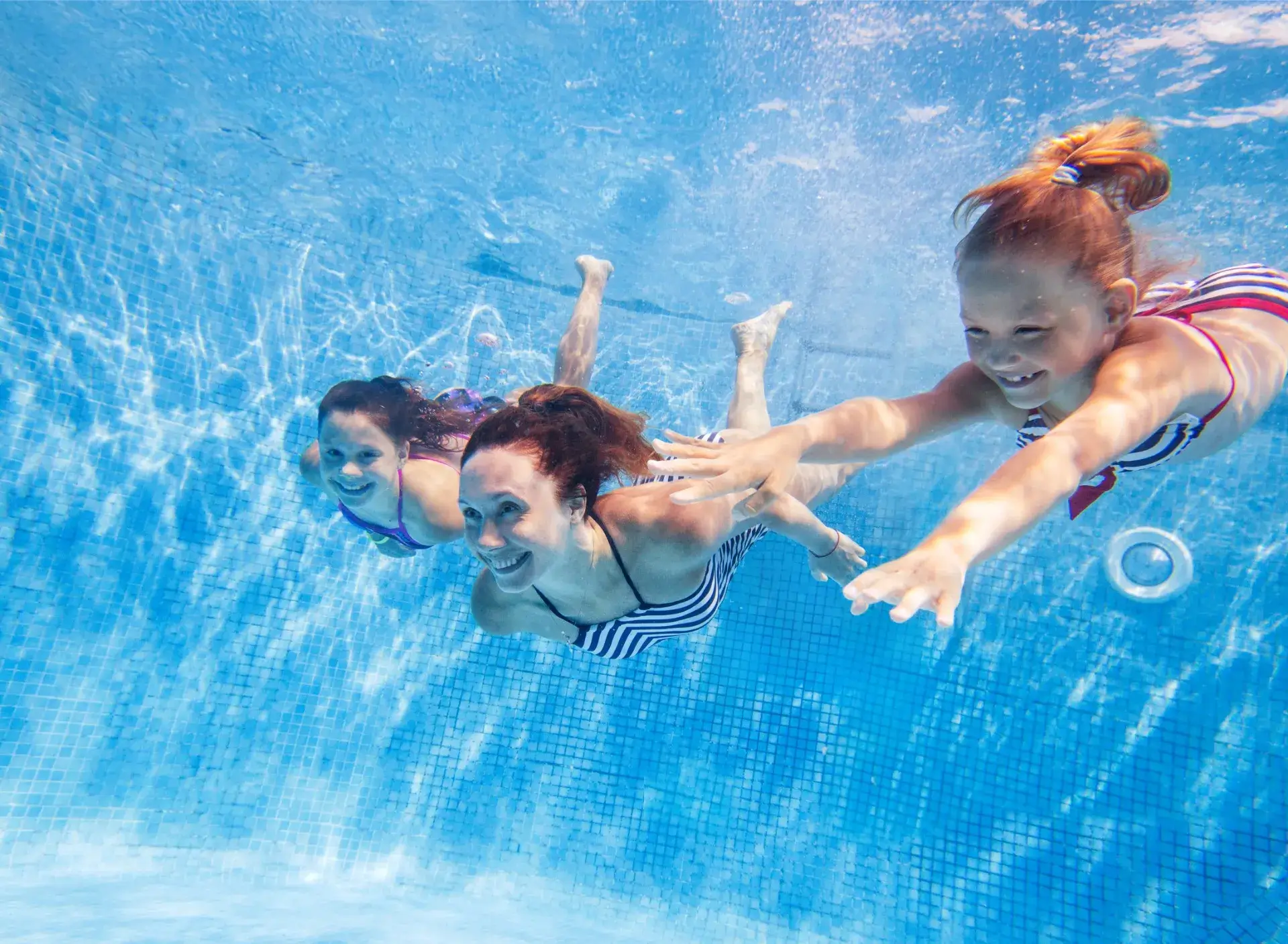 Woman and two girls swimming underwater in a pool, smiling and reaching forward.
