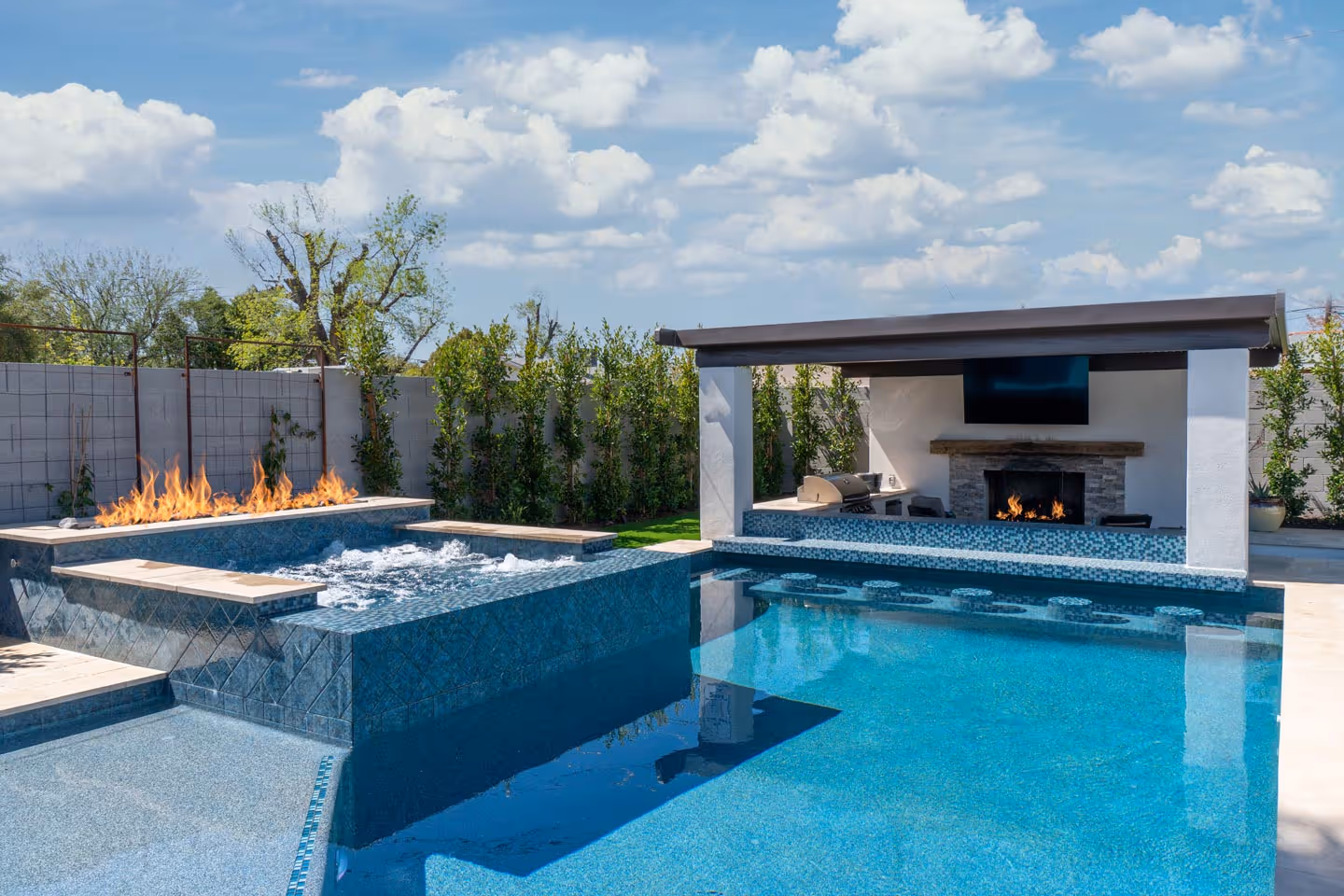 Modern backyard pool with built-in hot tub, fire feature, swim-up bar, outdoor fireplace, and a grill under a shaded pavilion.