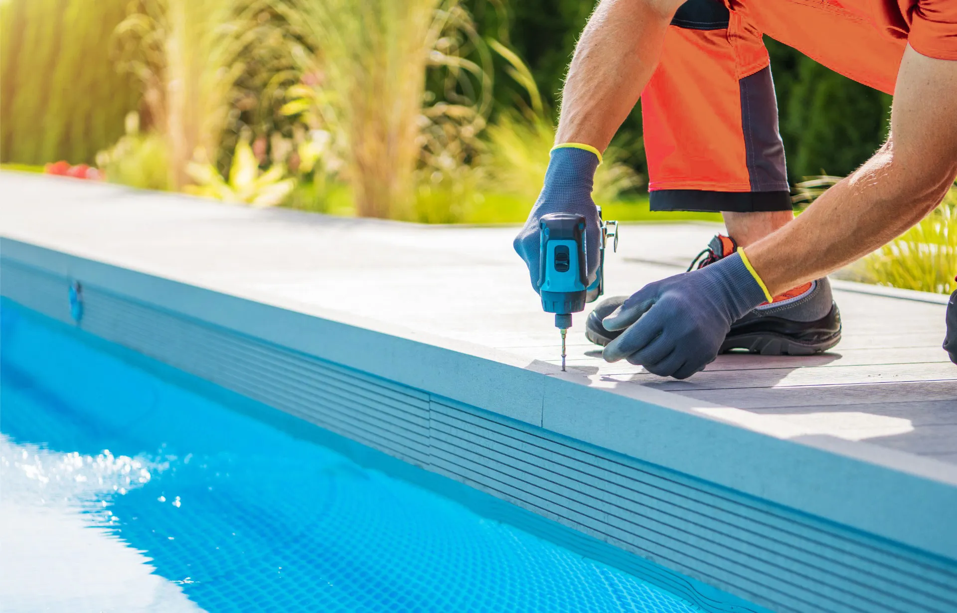 Person in orange shorts and blue gloves using a cordless drill to fix the edge of a poolside deck.